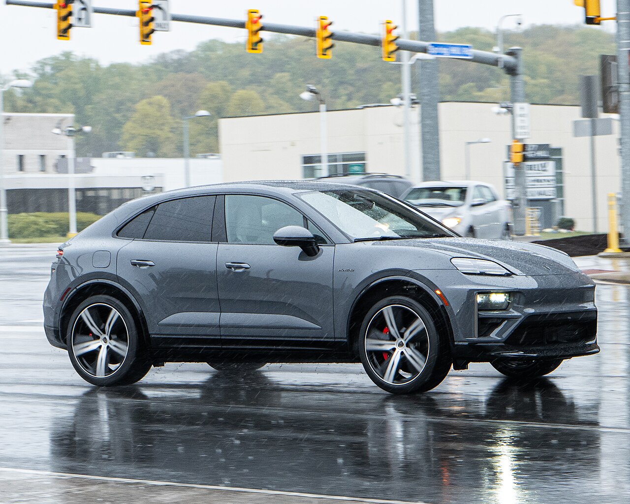 A grey Porsche Macan electric SUV driving on a wet road at an intersection.