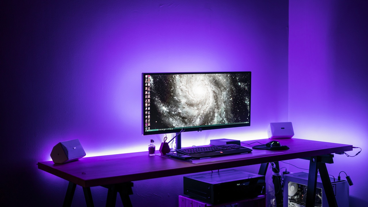 A clean gaming desk setup with a computer monitor displaying a galaxy, illuminated by purple ambient lighting.