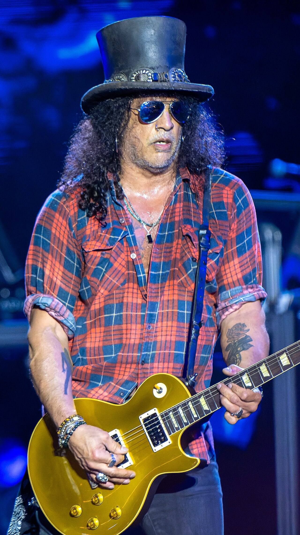 A portrait photo of the guitarist Slash playing his guitar on stage, wearing his signature top hat and sunglasses.