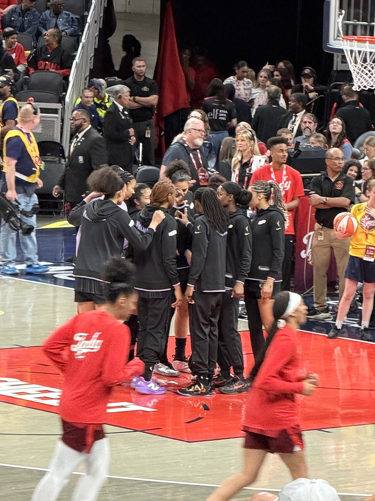 A group of female basketball players in black warm-up gear huddled together on a court before a game.